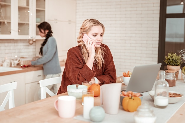 Vriend koken achter. Drukke vastberaden blond meisje in oranje oversized trui met telefoontje tijdens het ontbijt