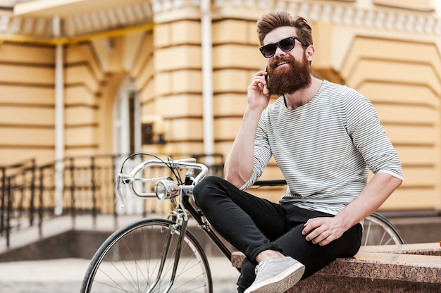 Vriend uitnodigen voor een ritje. gelukkige jonge, bebaarde man die op de mobiele telefoon praat en glimlacht terwijl hij buiten in de buurt van zijn fiets zit