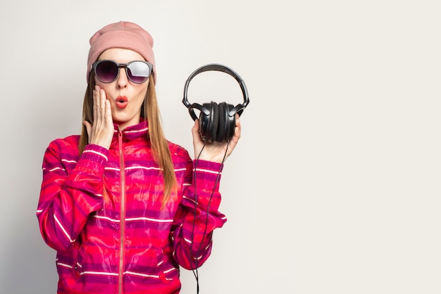 Vriendelijk emotioneel. Jonge vrouw met bril, een hoed en een roze sportjasje houdt een koptelefoon