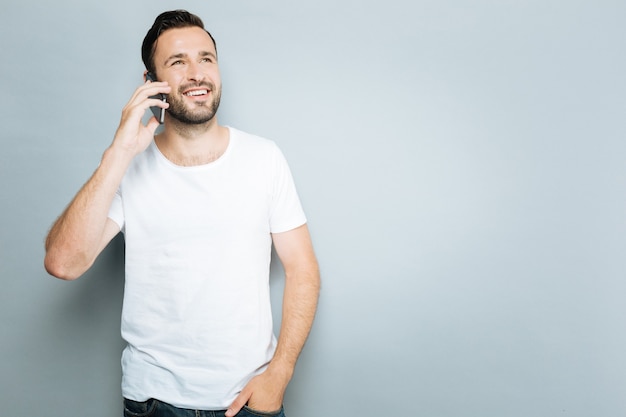 Vriendelijk gesprek. Glimlachende mannelijke bedrijfstelefoon dichtbij oor en linkerhand in zak steken, die zich tegen grijze achtergrond bevindt