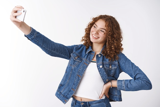 Vriendelijk uitgaande aantrekkelijke jonge roodharige krullend meisje puistjes en sproeten houden hand taille zelfverzekerd verlengen arm nemen selfie nieuwe smartphone camera glimlachend gadget display staande witte achtergrond