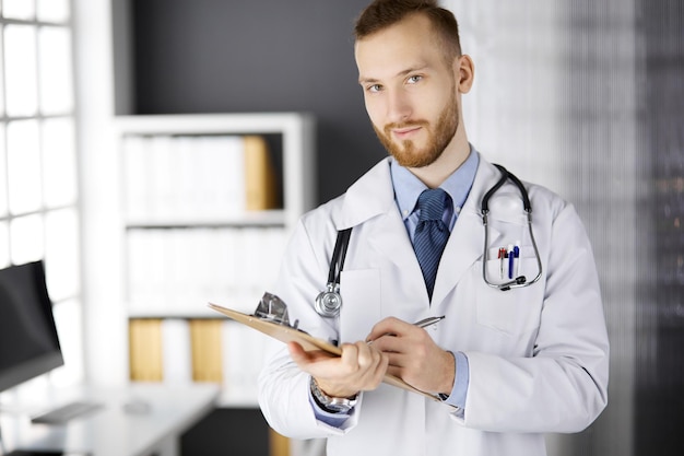 Vriendelijke arts met een rode baard die staat en schrijft met klembord in de kliniek op zijn werkplek. Perfecte medische service in het ziekenhuis. Geneeskunde, gezondheidszorg.