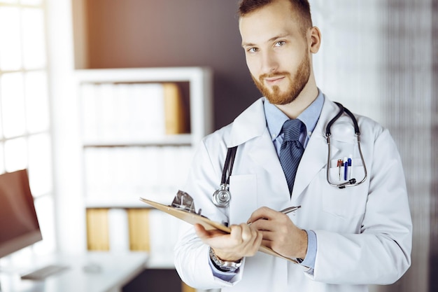 Vriendelijke arts met een rode baard die staat en schrijft met klembord in een zonnige kliniek op zijn werkplek. Geneeskunde concept.