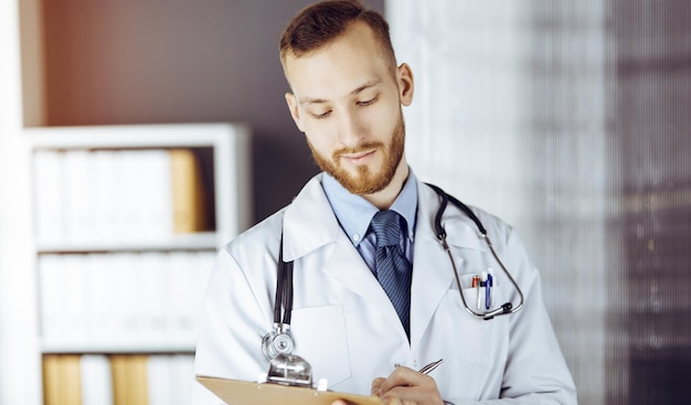 Vriendelijke arts met een rode baard die staat en schrijft met klembord in een zonnige kliniek op zijn werkplek. Geneeskunde concept.