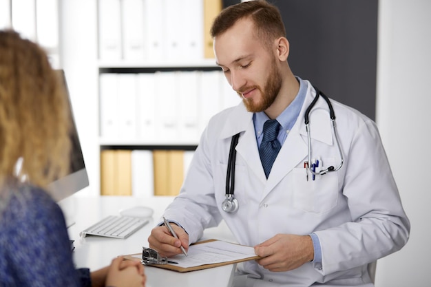 Vriendelijke arts met rode baard en geduldige vrouw bespreken het huidige gezondheidsonderzoek terwijl ze in de kliniek zitten.