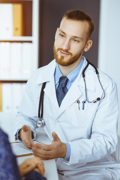 Vriendelijke arts met rode baard en geduldige vrouw bespreken het huidige gezondheidsonderzoek terwijl ze in een zonnige kliniek zitten. Geneeskunde concept.