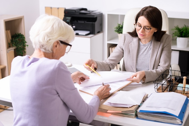 Vriendelijke financieel adviseur die met senior vrouw werkt en plaats toont voor ondertekening in document