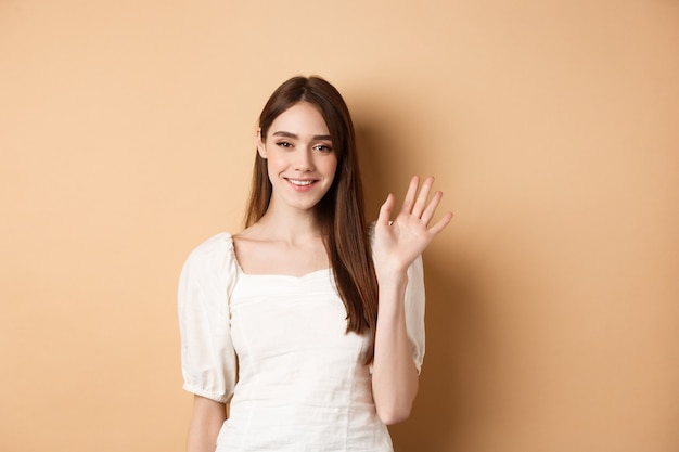 Vriendelijke glimlachende vrouw zegt hallo, hand afwijzend om u te begroeten, vrolijk permanent op beige achtergrond.