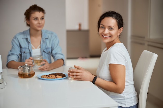 Vriendelijke internationale vrouw die de beker in beide handen houdt terwijl ze geniet van gezellige communicatie met haar vriend