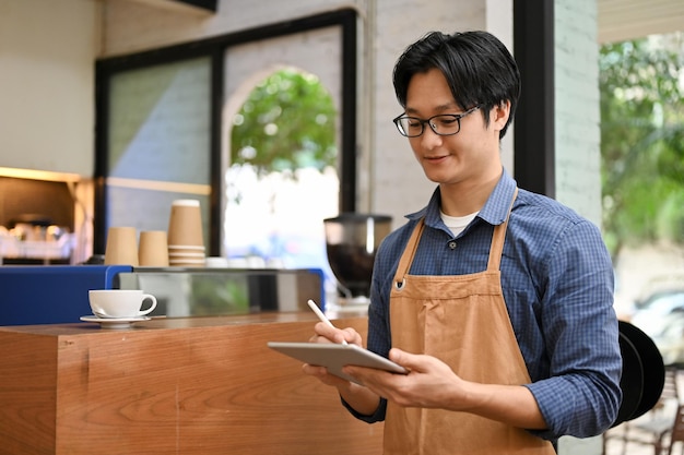 Vriendelijke jonge Aziatische mannelijke ober of barista in schort met behulp van digitale tablet die drankbestellingen opmerkt