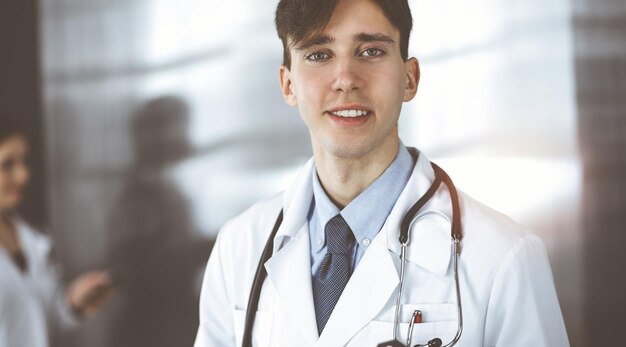 Vriendelijke jonge mannelijke arts die met een klembord staat in een moderne kliniek Vrouwelijke collega is bezig met het invullen van medicatie dossiers formulier op de achtergrond van de arts Geneeskunde concept