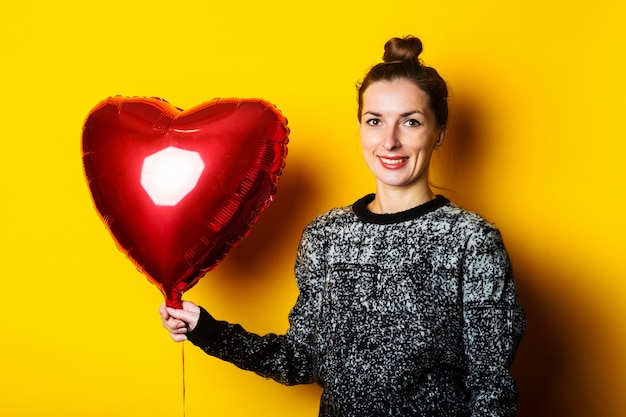 Vriendelijke jonge vrouw met een rode luchtballon in de vorm van een hart op een gele achtergrond.