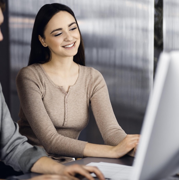 Vriendelijke jonge zakenman en programmeur in een groen shirt werkt aan een computer, terwijl ze samen aan het bureau zitten met een vrouwelijke collega in een modern kantoor.