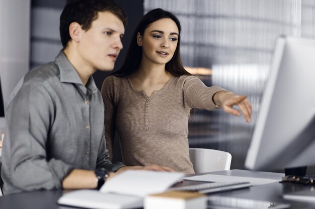 Vriendelijke jonge zakenman en programmeur in een groen shirt werkt op de computer, terwijl hij samen aan het bureau zit met een vrouwelijke collega in een modern kantoor. Focus op vrouw. Concept van succesvol