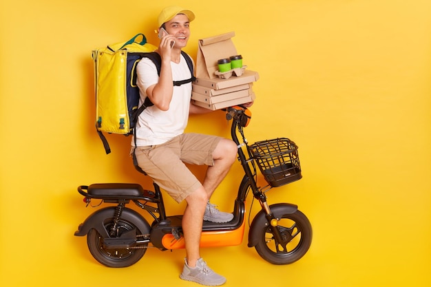 Vriendelijke koerier op elektrische fiets met thermorugzak die pizza en koffie levert om op mobiele telefoon te praten en naar de camera te kijken met een glimlach geïsoleerd op gele achtergrond