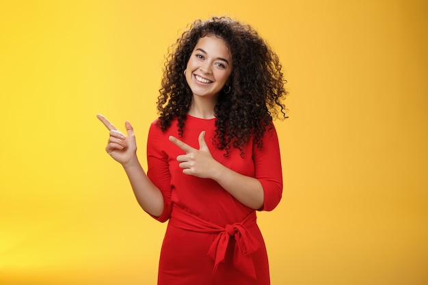 Vriendelijke schattige assertieve vrouwelijke assistent in elegante rode jurk met krullend haar kantelend hoofd flirterig en ...