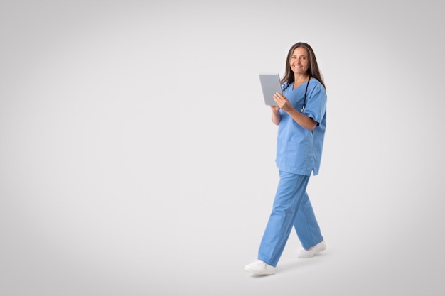 Vriendelijke senior vrouw in medisch uniform met digitale tablet lopend op grijze achtergrond vol