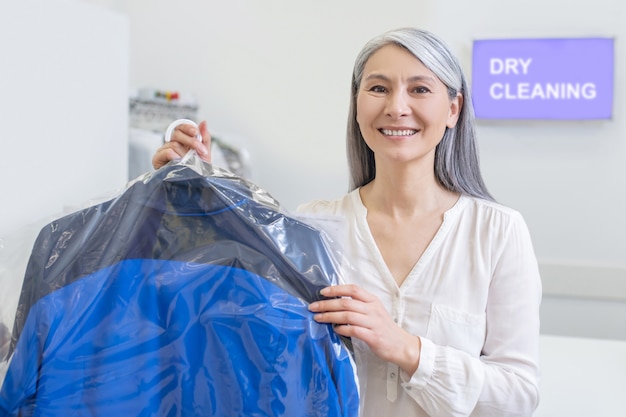 Vriendelijke vrolijke vrouw met lang grijs haar met schone ingepakte kleren in hand in de stomerij