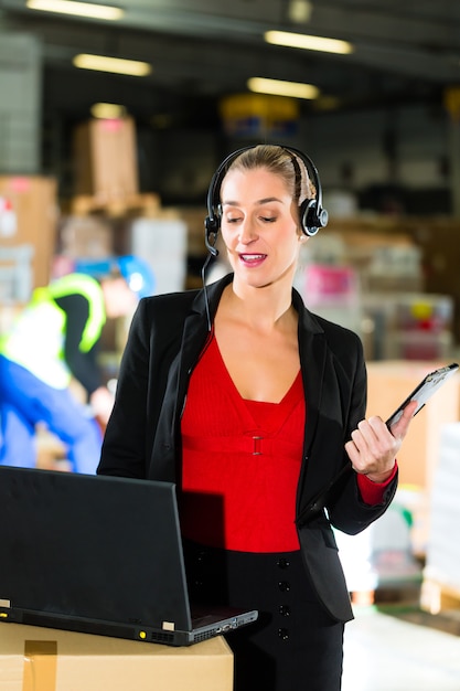 Vriendelijke vrouw, coördinator of supervisor met behulp van headset en laptop in magazijn van expeditiebedrijf,