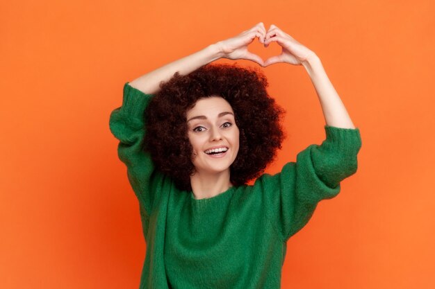 Vriendelijke vrouw met Afro-kapsel, gekleed in een groene casual stijl trui met opgeheven armen en hartvorm met vingers, kijkend naar de camera. Indoor studio opname geïsoleerd op een oranje achtergrond.