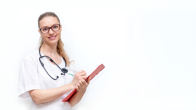 Vriendelijke vrouwelijke arts in witte jas met phonendoscope maakt aantekeningen op klembord geïsoleerd op een witte achtergrond medische concept van jonge mooie vrouw ziekenhuis werknemer camera kijken en glimlachen