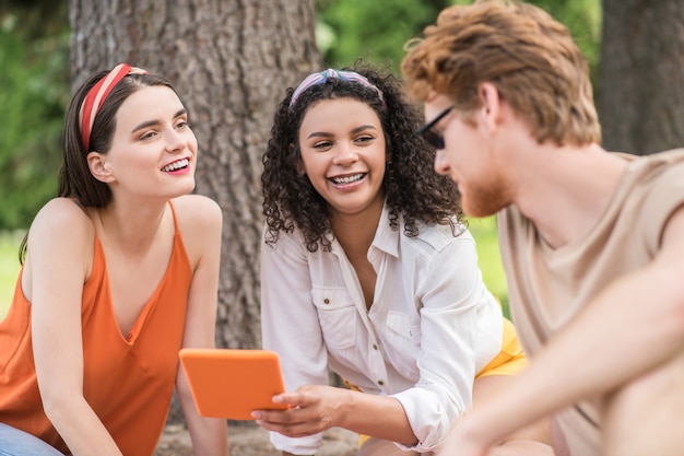 Vrienden, communicatie. Man in zonnebril en twee lachende vriendinnen met tablet vriendelijk chatten op picknick in park