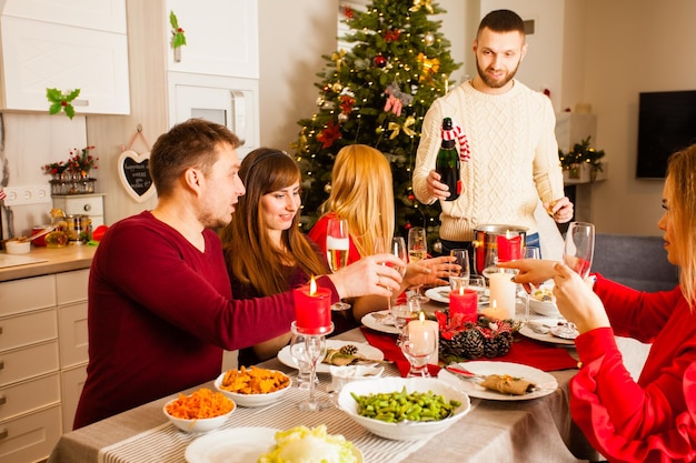 Vrienden die kerst of oud en nieuw vieren, zittend in een stijlvolle eetkamer rond de tafel met veel gerechten, brandende kaarsen. Man staande met fles champagne die het in glazen giet