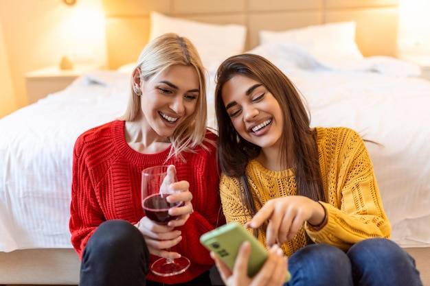 Vrienden met wijn op de vloer thuis Gelukkig lachende jonge vrouwen vrienden kijken naar foto's op mobiele telefoon Meisjes hebben plezier Twee vrouwelijke vrienden ontspannen thuis met een glas wijn samen praten