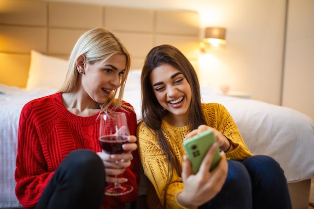 Vrienden met wijn op de vloer thuis Gelukkige glimlachende jonge vrouwenvrienden die foto's bekijken op mobiele telefoon