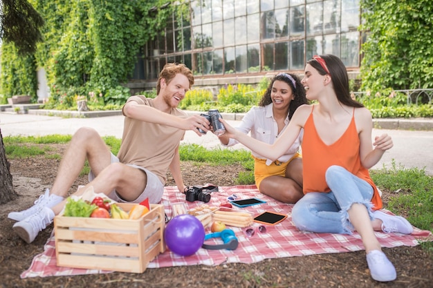 Vrienden, toast. Jonge vrolijke mensen zitten met een drankje in uitgestrekte hand naar elkaar op plaid in park op zonnige dag