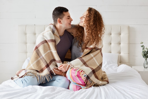 vriendin en vriend dragen nonchalant gekleed, genieten van gezelligheid onder plaid in slaapkamer, omhelzen elkaar, kijken in de ogen en praten gezellig. Net getrouwd verliefde paar samen tijd doorbrengen.