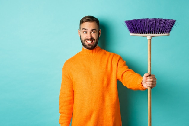 Vriendje kijkt verward naar bezem, dwingt huis schoon, doet klusjes, staat ontevreden tegen lichtblauwe achtergrond.