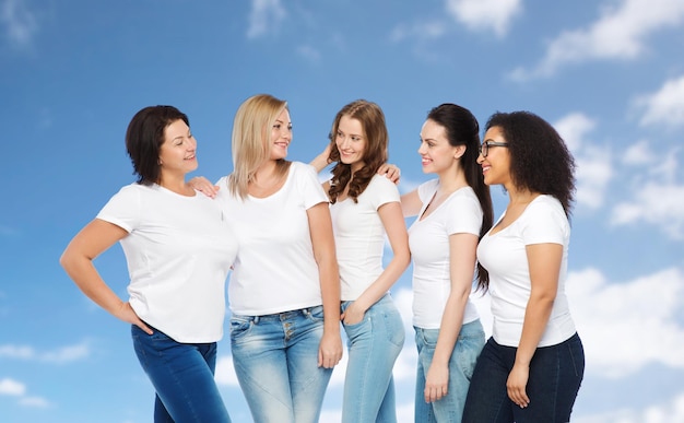 Foto vriendschap, divers, lichaam positief en mensen concept - groep gelukkige vrouwen van verschillende grootte in witte t-shirts knuffelen over blauwe lucht en wolken achtergrond