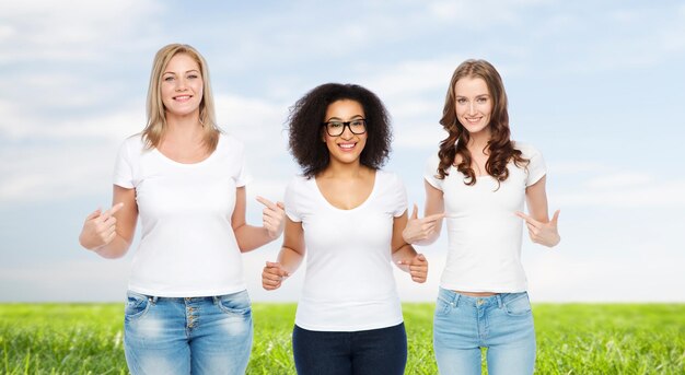 vriendschap, divers, lichaamspositief en mensenconcept - groep gelukkige vrouwen van verschillende grootte in witte t-shirts die met de vinger naar zichzelf wijzen over blauwe lucht en grasachtergrond