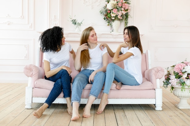 Vriendschap en geluk concept - drie vriendinnen praten thuis. Drie gelukkige, grappige multi-etnische dames, beste vrienden die lachen terwijl ze plezier hebben, mooie diverse vrouwen dragen witte t-shirts en jeans