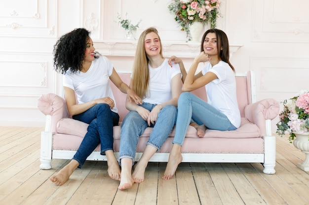 Vriendschap en geluk concept - drie vriendinnen praten thuis. Drie gelukkige, grappige multi-etnische dames, beste vrienden die lachen terwijl ze plezier hebben, mooie diverse vrouwen dragen witte t-shirts en jeans