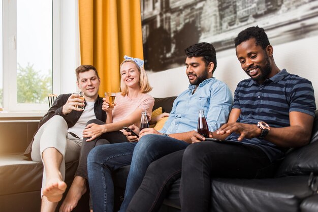 Vriendschap, entertainmentconcept. Schot van vier gelukkige vrienden die op de bank zitten en glimlachen terwijl ze tv kijken en koolzuurhoudende dranken en sap drinken, thuis plezier hebben met een modern interieur.
