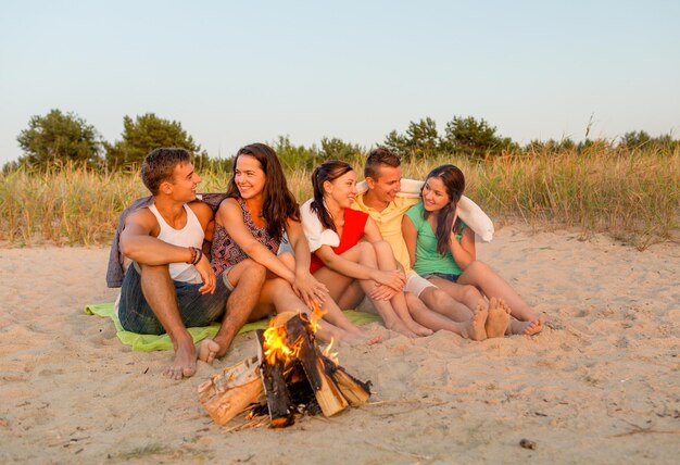 Vriendschap, geluk, zomervakantie, vakantie en mensenconcept - groep glimlachende vrienden die dichtbij brand op strand zitten