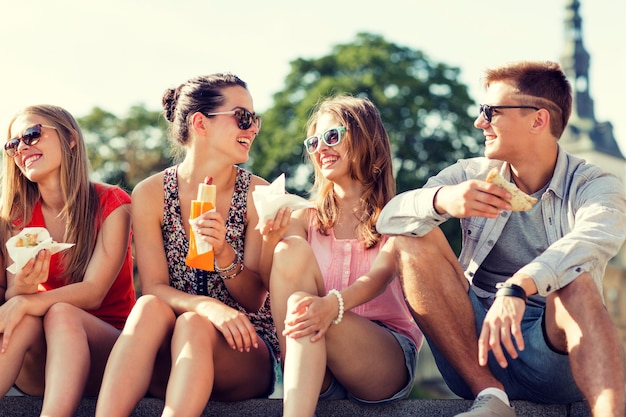 vriendschap, vrije tijd, zomer en mensen concept - groep lachende vrienden in zonnebril zittend met eten op stadsplein