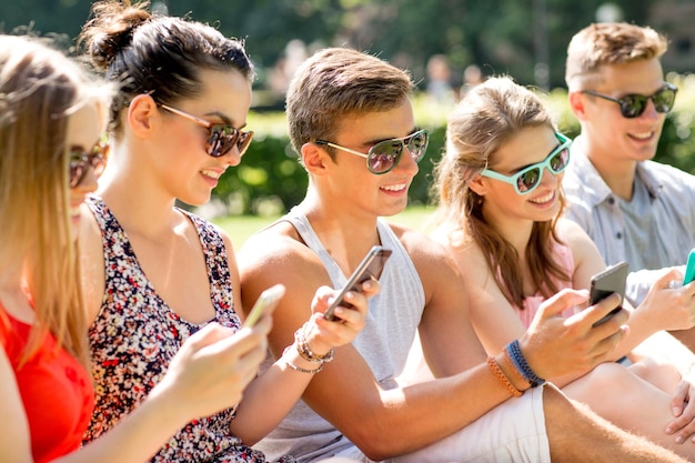 vriendschap, vrije tijd, zomer, technologie en mensenconcept - groep glimlachende vrienden met smartphones die op gras in park zitten