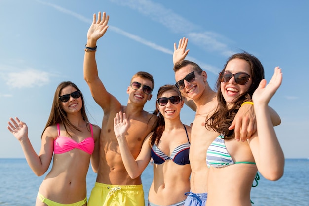 vriendschap, zee, vakantie, gebaar en mensenconcept - groep glimlachende vrienden die zwemkleding en zonnebril dragen die handen op strand zwaaien