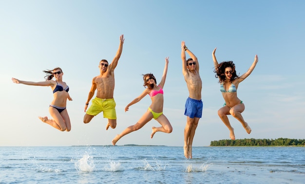 vriendschap, zee, zomervakantie, vakantie en mensenconcept - groep lachende vrienden die badkleding en zonnebril dragen die op strand springen