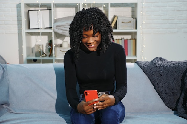 Vrij Afro-Amerikaanse dame met krullende haartypes op moderne rode smartphone zittend op een grijze bank in de kamer