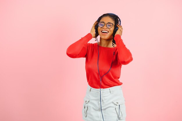 Vrij Afro-Amerikaanse jonge vrouw met heldere glimlach gekleed in casual kleding, bril en koptelefoon dansen over roze achtergrond.