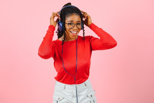 Vrij Afro-Amerikaanse jonge vrouw met heldere glimlach gekleed in casual kleding, bril en koptelefoon dansen over roze achtergrond.