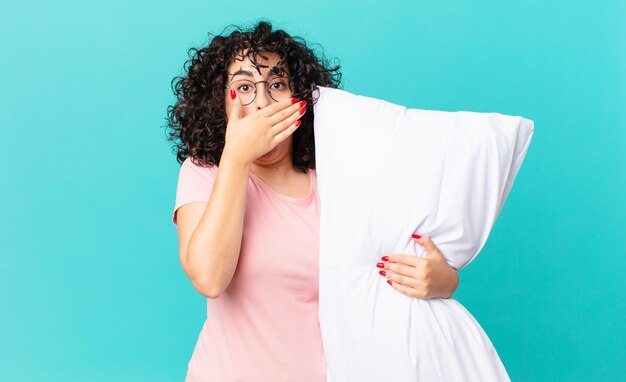 Vrij arabische vrouw die mond bedekt met handen met een geschokt. een pyjama dragen en een kussen vasthouden