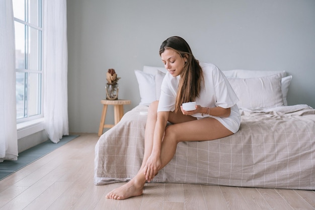 Vrij blanke jonge volwassen Italiaanse vrouw zit op bed en doet bodylotion op tevreden benen