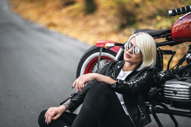 Vrij blond biker meisje in zonnebril zittend in de buurt van rode motorfiets op de weg in het bos