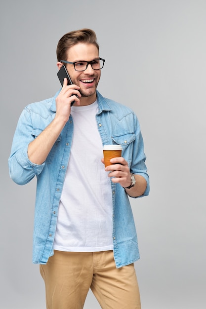 Vrij casual man in spijkerbroek shirt met zijn telefoon en kopje koffie te gaan staan over studio grijze muur