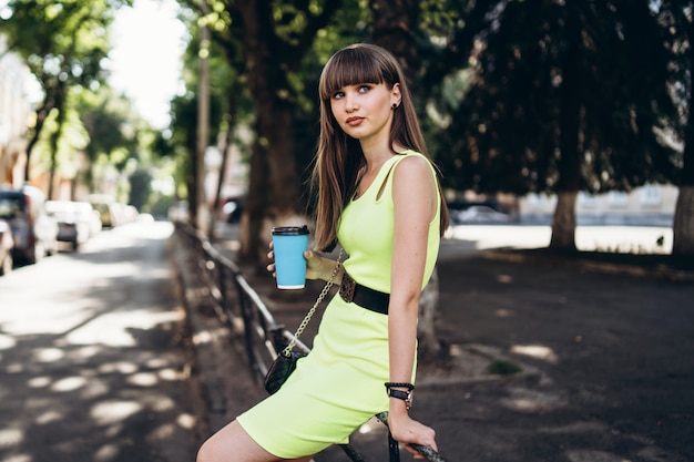 Vrij donkerbruin meisje in groene kleding met kop van koffie lopen openlucht op de straat